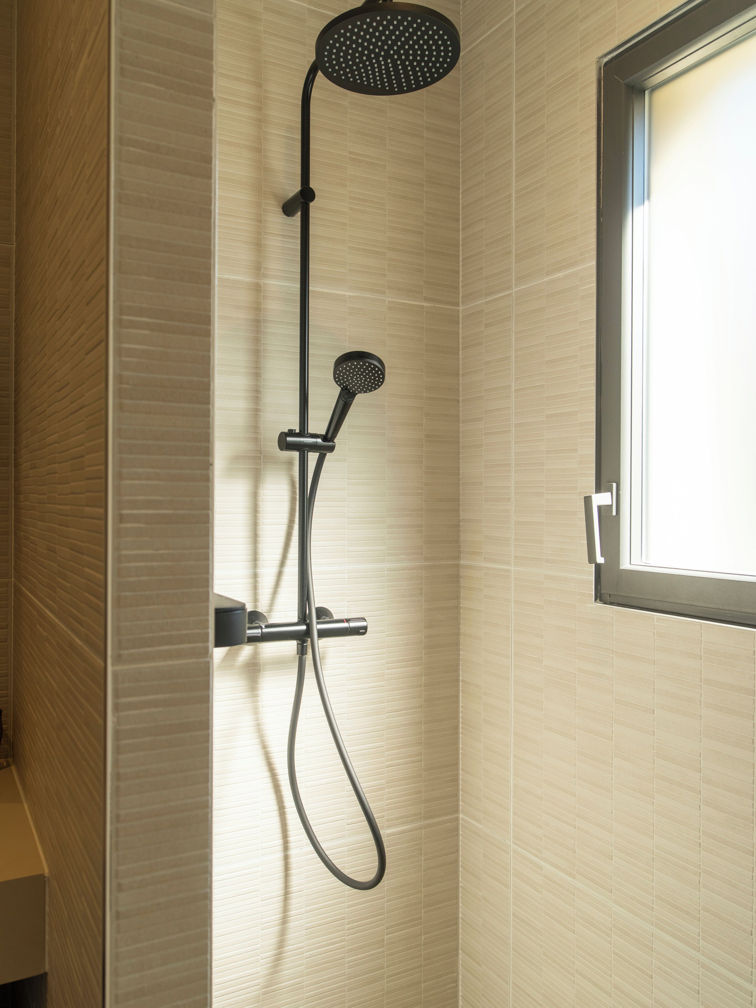 A shower with a handheld showerhead and a fixed showerhead, a window, and a tiled wall.