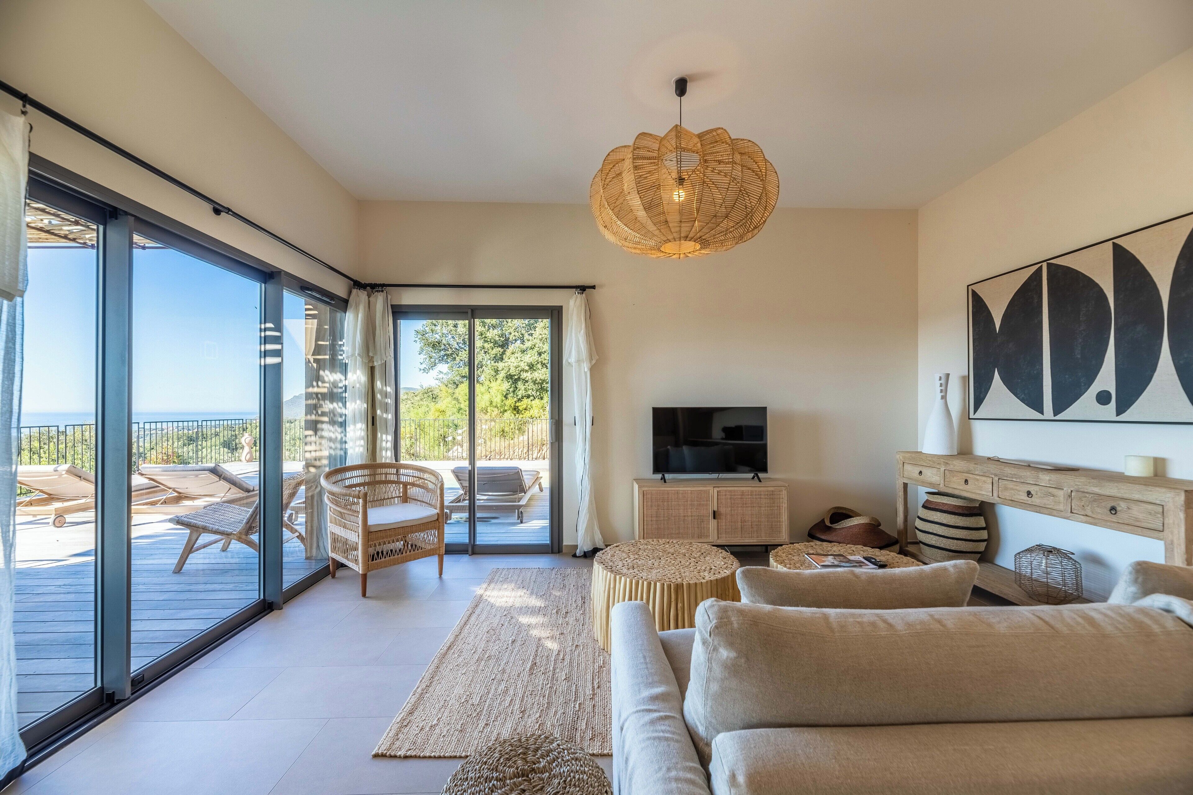 A modern living room with a sofa, a television, a wicker lamp, and a view of the outdoors through large glass doors.