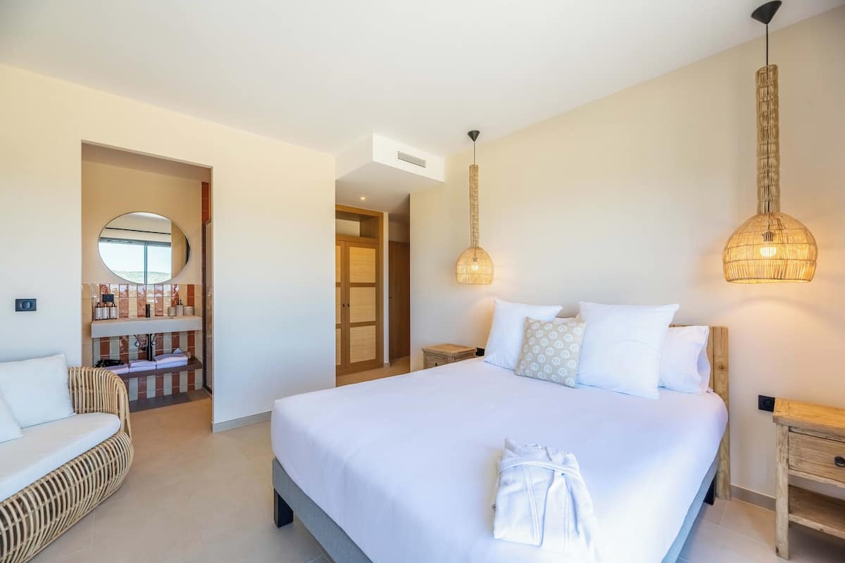 Ein Hotelzimmer mit einem großen Bett, einem runden Spiegel, einer Korbschaukel, einem Holznachttisch und einer Hängeleuchte.