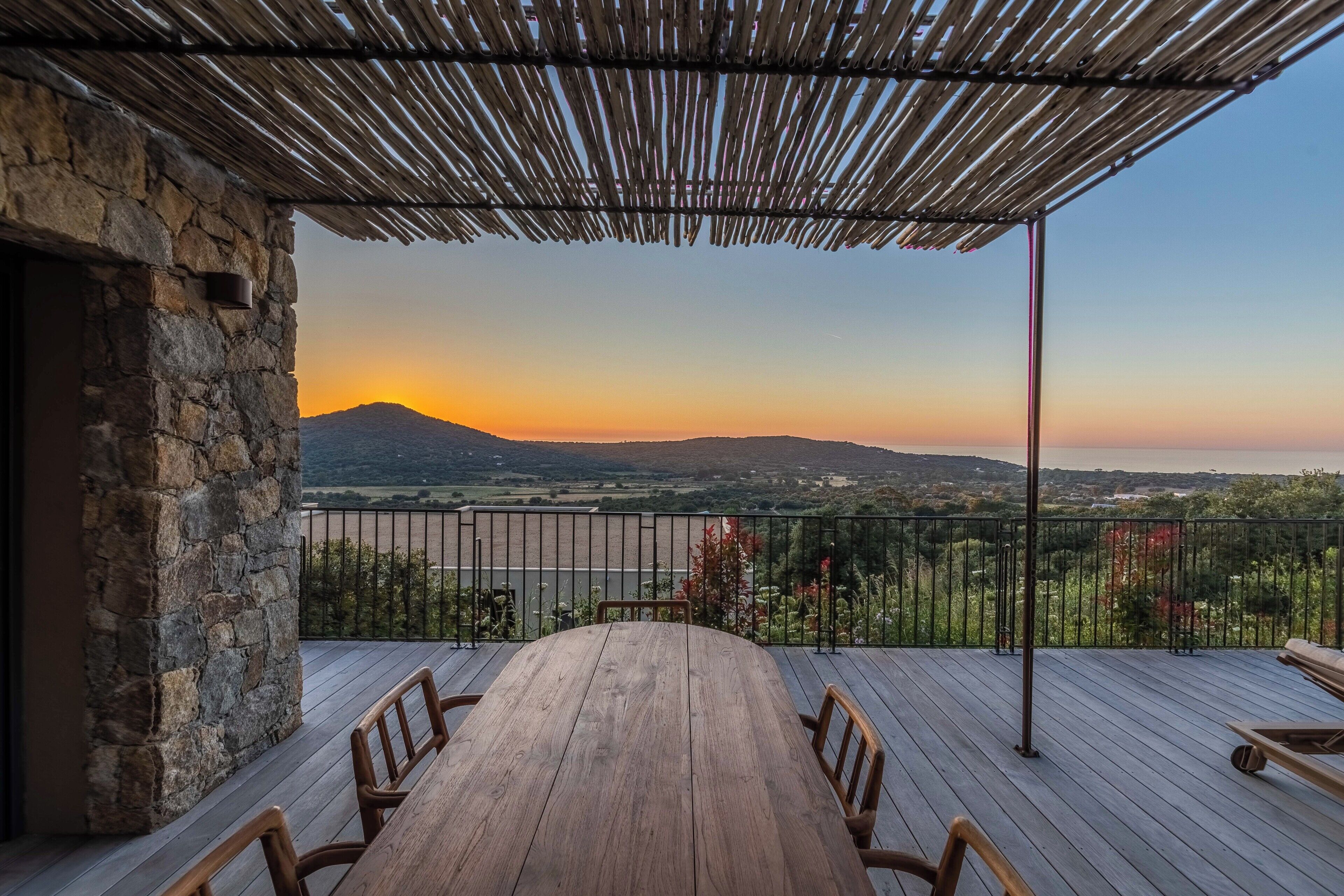 A wooden deck with a table and chairs, a stone wall, and a view of a landscape with a mountain and sunset.