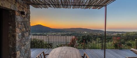 A wooden deck with a table and chairs, a stone wall, and a view of a landscape with a mountain and sunset.