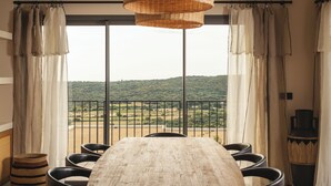 A dining area with a large wooden table and black chairs, a wicker pendant light, and a view of a green landscape through large windows. -  Résidence & Villas Oradoro (Belgodere)