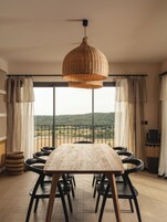 Un espace repas avec une grande table en bois et des chaises noires, un luminaire suspendu en osier, et une vue sur un paysage verdoyant grâce à de grandes fenêtres.