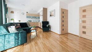 Una sala de estar moderna con sofá, sillón y mesa de centro. Se observa una cocina al fondo con armarios y un lavamanos.
