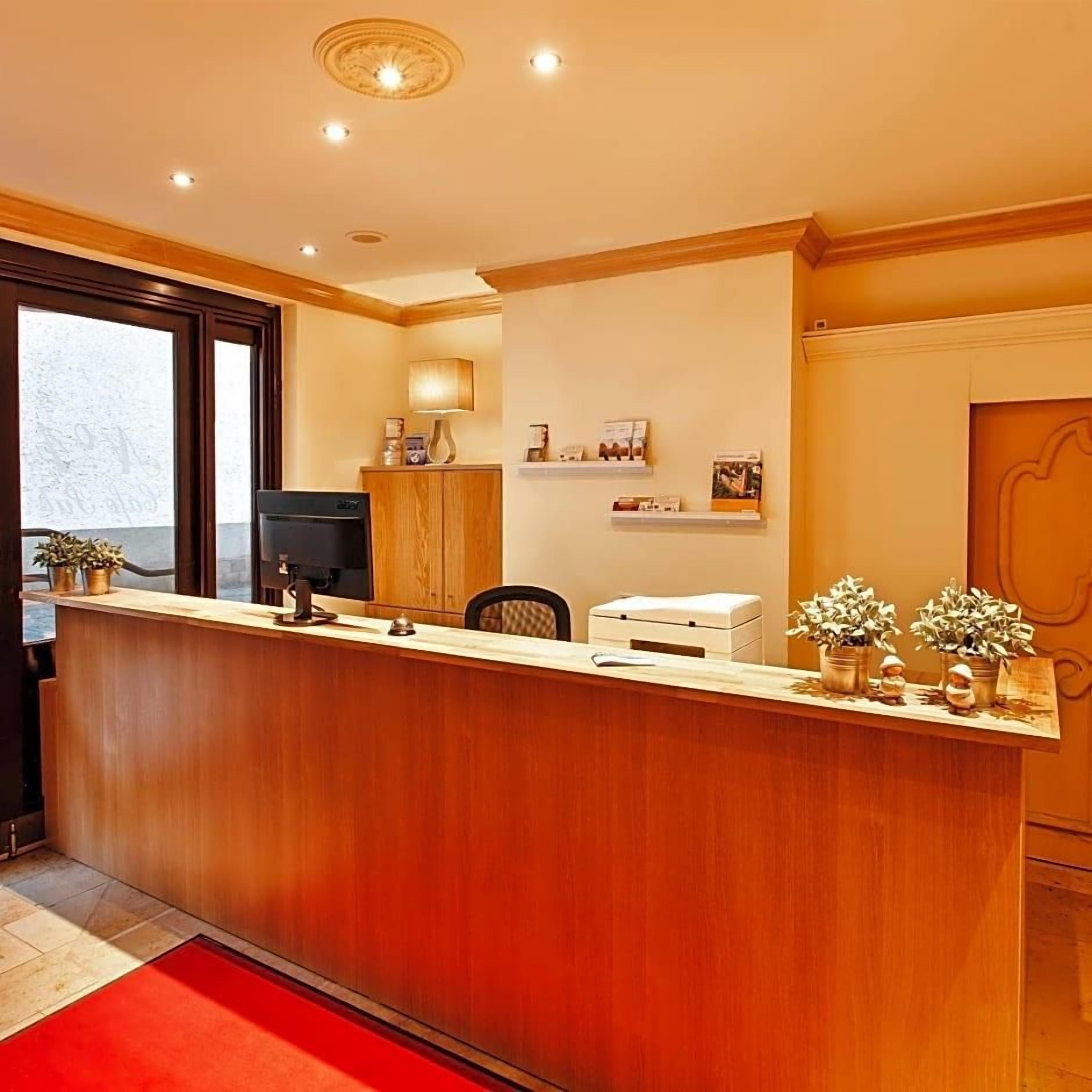 A reception desk with a flat-screen TV, a chair, and a desk lamp. There are potted plants and a red rug on the floor.