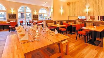 A restaurant with wooden tables and chairs, large windows, and wall-mounted lights.