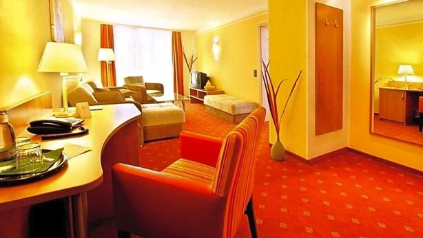 Quarto de hotel com carpete vermelho, escrivaninha com abajur, sofá, televisão e espelho.