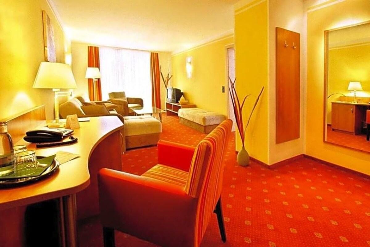 Habitación de hotel con alfombra roja, un escritorio con lámpara, un sofá, un televisor y un espejo.