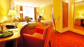 A hotel room with a red carpet, a desk with a lamp, a sofa, a TV, and a mirror.