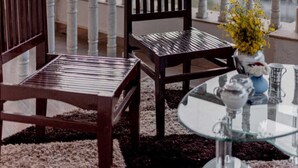 Un balcón con dos sillas de madera, una mesa de vidrio y un jarrón con flores amarillas.