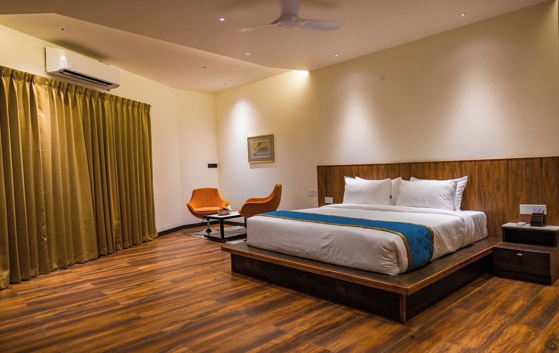 Une chambre d’hôtel avec un plancher en bois, un lit avec une literie blanche et un détail bleu, une chaise orange, une table de chevet en bois et un ventilateur de plafond.