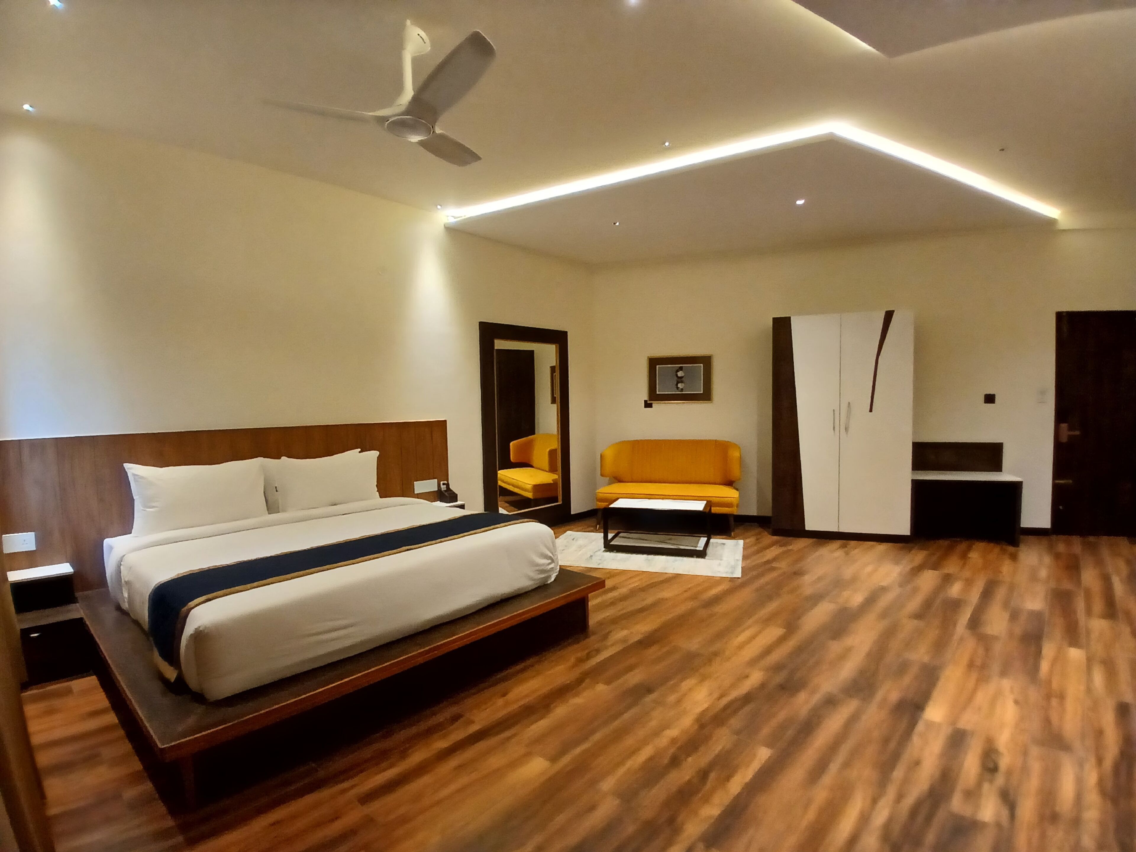 A modern hotel room with a wooden floor, a bed with white linens, a ceiling fan, a sofa, a wardrobe, and a small table.
