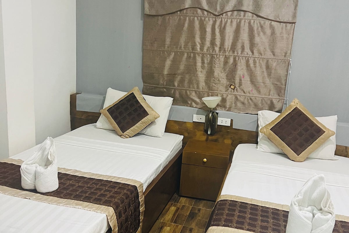 A hotel room with two single beds, wooden flooring, a headboard with a curtain, a bedside table with a lamp, and a wall-mounted air conditioner.