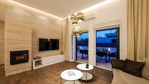 Una sala moderna con una televisión de pantalla plana, chimenea, dos mesas redondas y un amplio ventanal corredero que da a un patio exterior.
