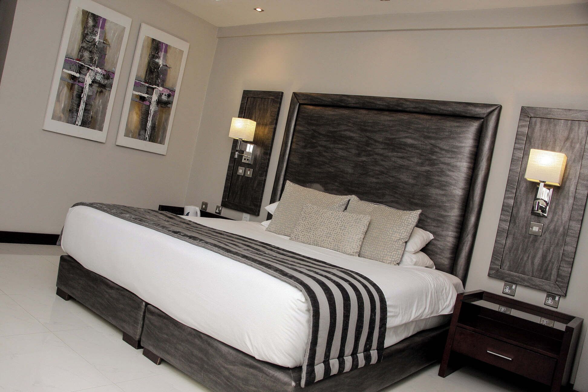 A modern bedroom with a large bed, dark headboard, bedside lamps, and artwork on the walls.