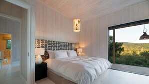 Una camera da letto con una testiera effetto pietra, un letto grande, due lampade ai comodini e vista su una foresta attraverso una porta scorrevole in vetro.