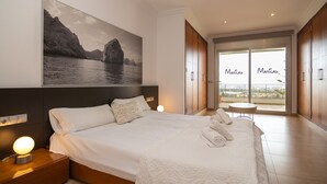 A modern hotel room with a large bed, a nightstand with a lamp, a painting of a coastal landscape, and a balcony with a view.