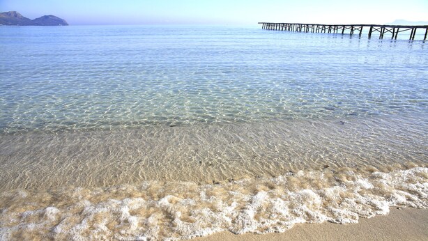 A beach with clear shallow waters, a sandy shore, and a pier extending into the sea.