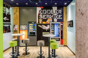 A modern bar area with stools, a 'COOLER SERVICE' sign, and vending machines. - B&B Hotel Köln-porz (Cologne)