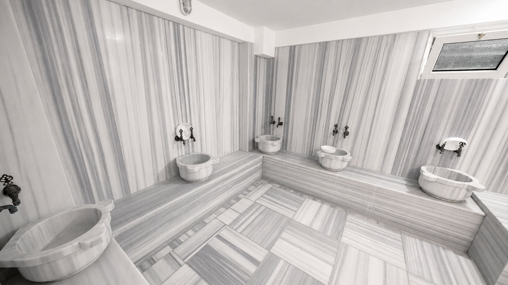 Un baño con paredes de mármol, varios lavamanos y un piso con diseño de espiga.