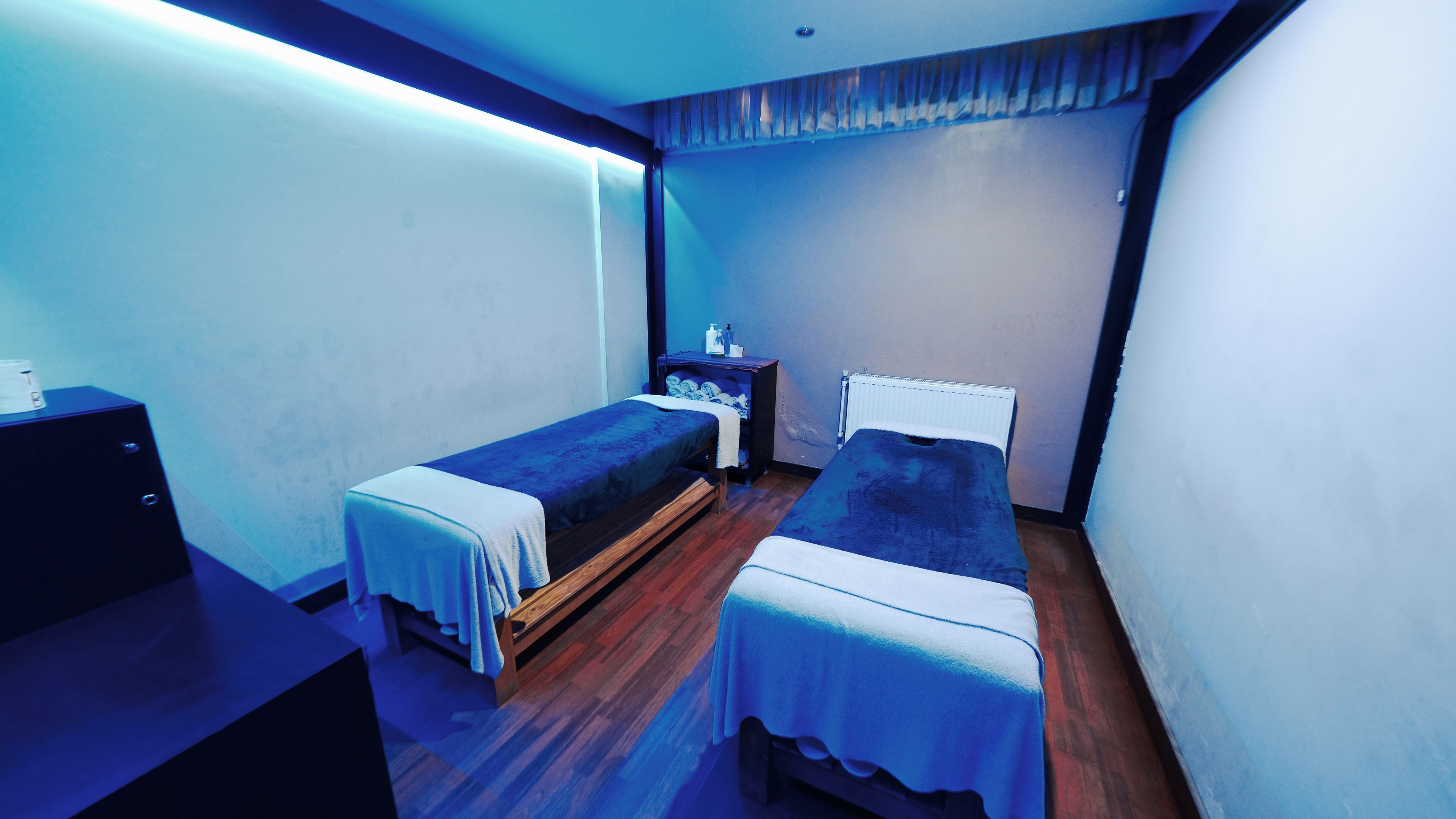 A small, dimly lit room with two single beds, a wooden dresser, and a nightstand with toiletries.
