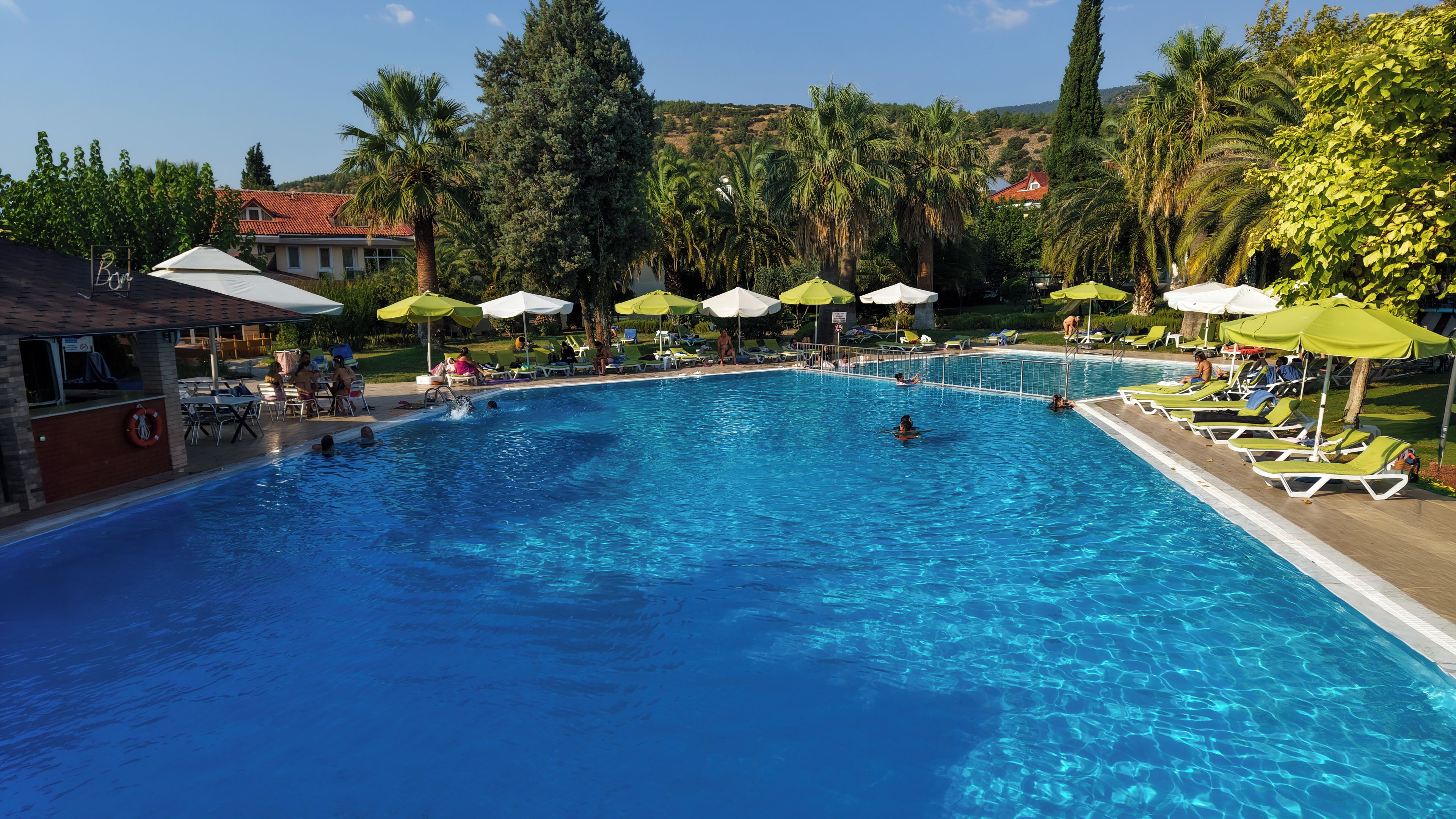 A swimming pool with clear blue water, surrounded by lounge chairs and umbrellas, with a building and trees in the background.