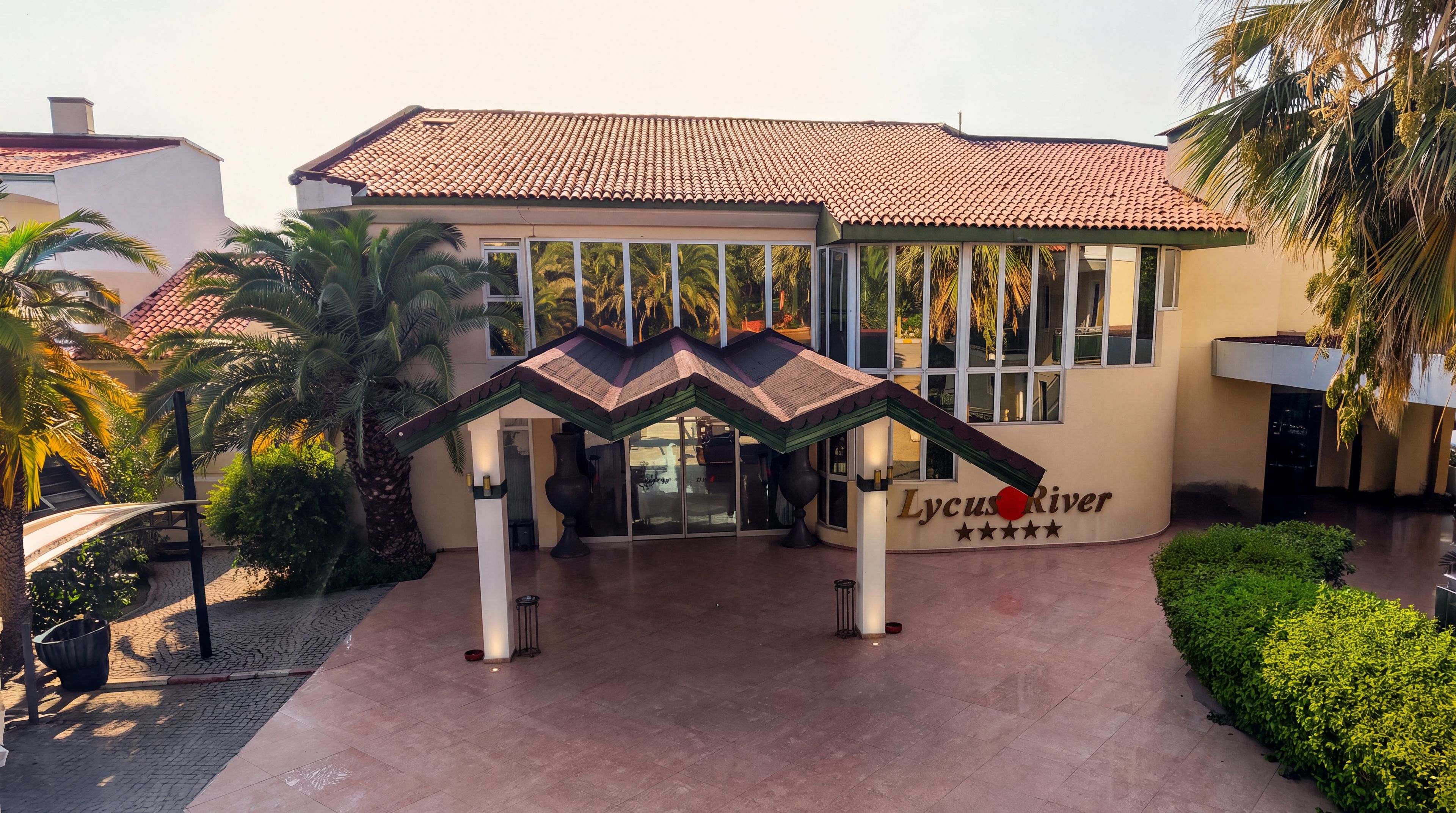 A hotel entrance with a tiled roof, a sign reading 'Lycus River,' and a palm tree in the foreground.