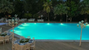 Zona junto a la piscina con mesas y sillas, rodeada de árboles y una piscina azul cristalina.