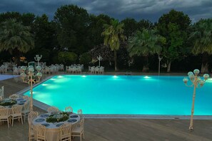 Zona junto a la piscina con mesas y sillas, rodeada de árboles y una piscina azul cristalina.