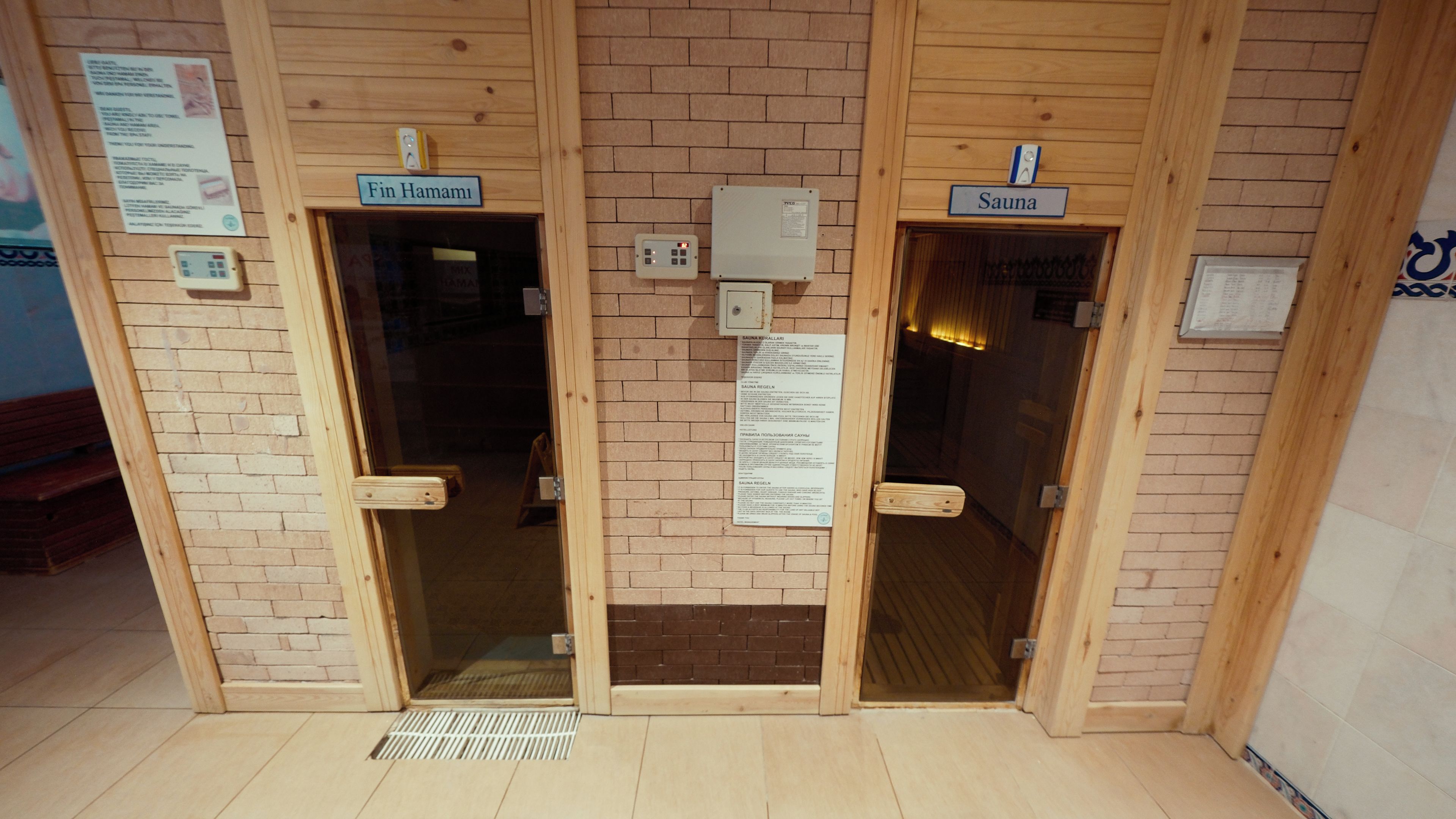 Tres puertas de madera con los rótulos 'Fin Hamami', 'Sauna' y 'Sauna', y una rejilla de ventilación en frente.