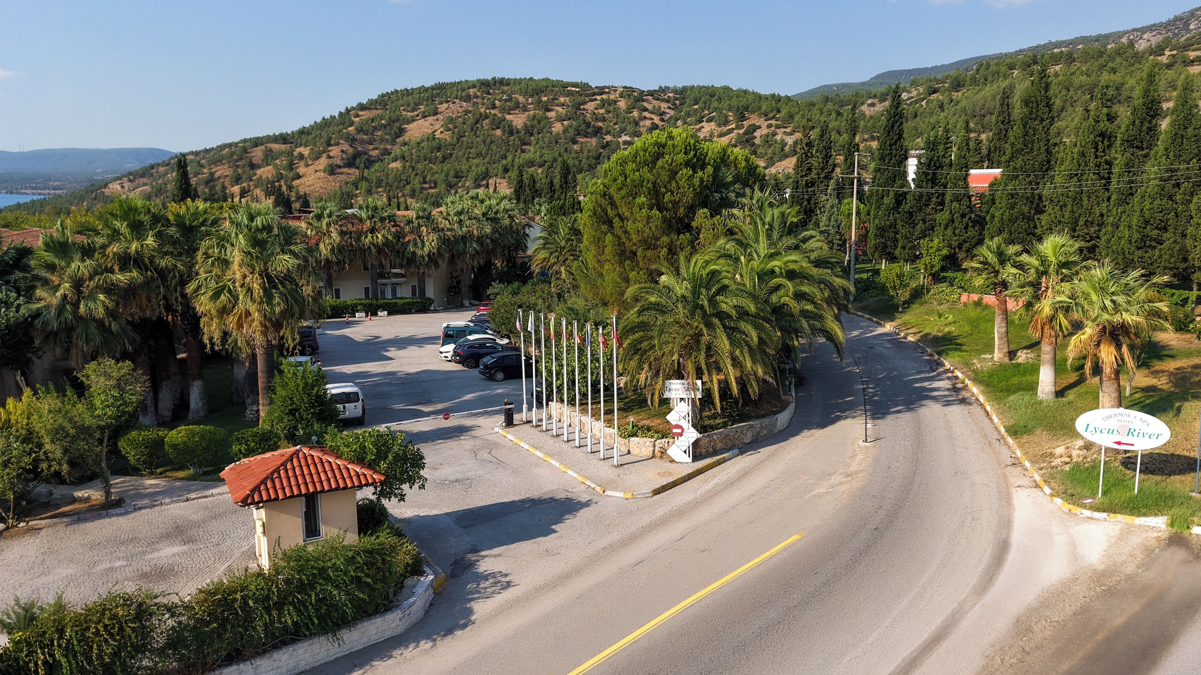 A coastal road with a hotel named 'Lycius River' on the right, surrounded by palm trees and a hilly backdrop.