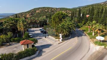 A coastal road with a hotel named 'Lycius River' on the right, surrounded by palm trees and a hilly backdrop.