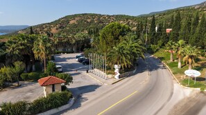 Una carretera costera con un hotel llamado 'Lycius River' a la derecha, rodeado de palmeras y un paisaje montañoso al fondo.