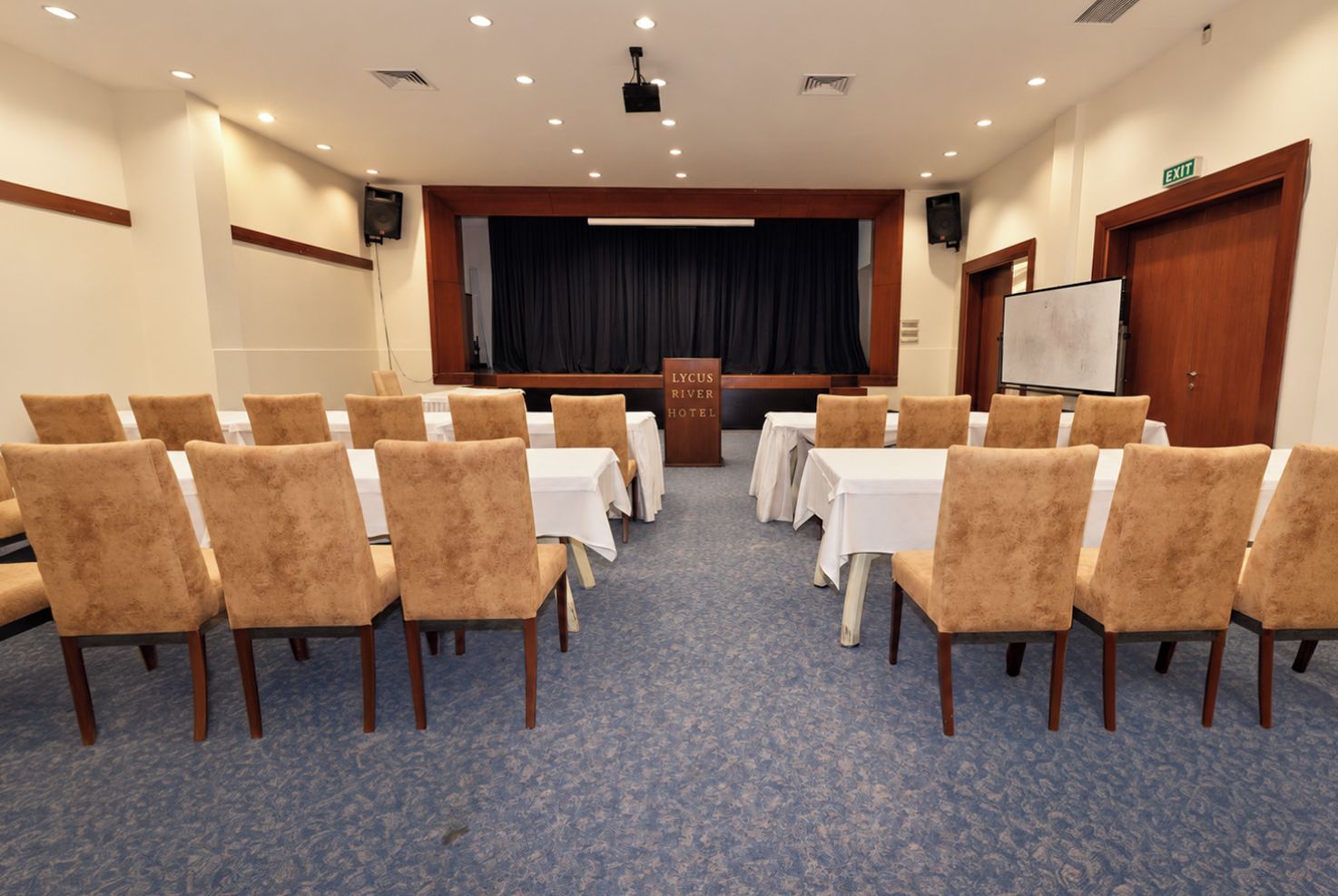 Un salón de conferencias con filas de sillas mirando hacia un escenario, una pizarra y una señal de salida.