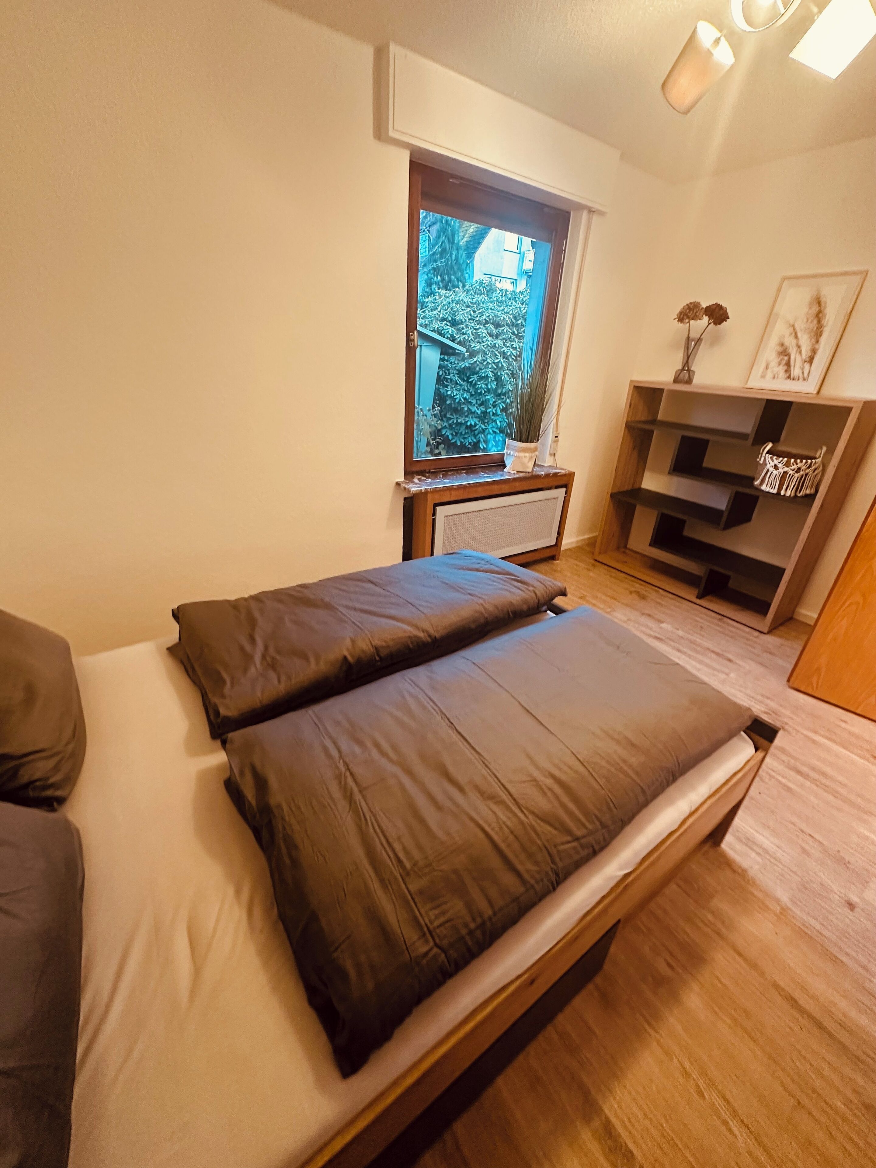 Una camera da letto con un letto, una finestra, un termosifone e una mensola in legno con oggetti decorativi.
