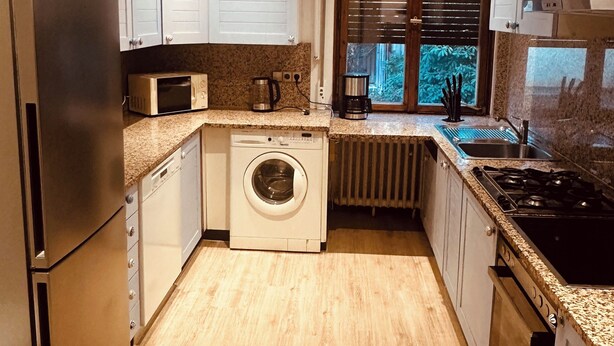 Una cocina moderna con electrodomésticos de acero inoxidable, armarios blancos, lavadora integrada y una ventana con persianas.