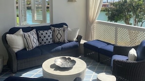 Uma área de estar coberta ao ar livre, com um sofá, cadeiras, uma mesa e vista para uma piscina e palmeiras.