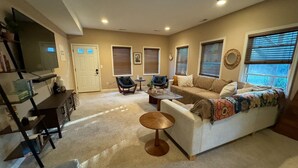 A living room with a sofa, armchairs, a coffee table, and a TV. There are windows with blinds, a ceiling fan, and a door.