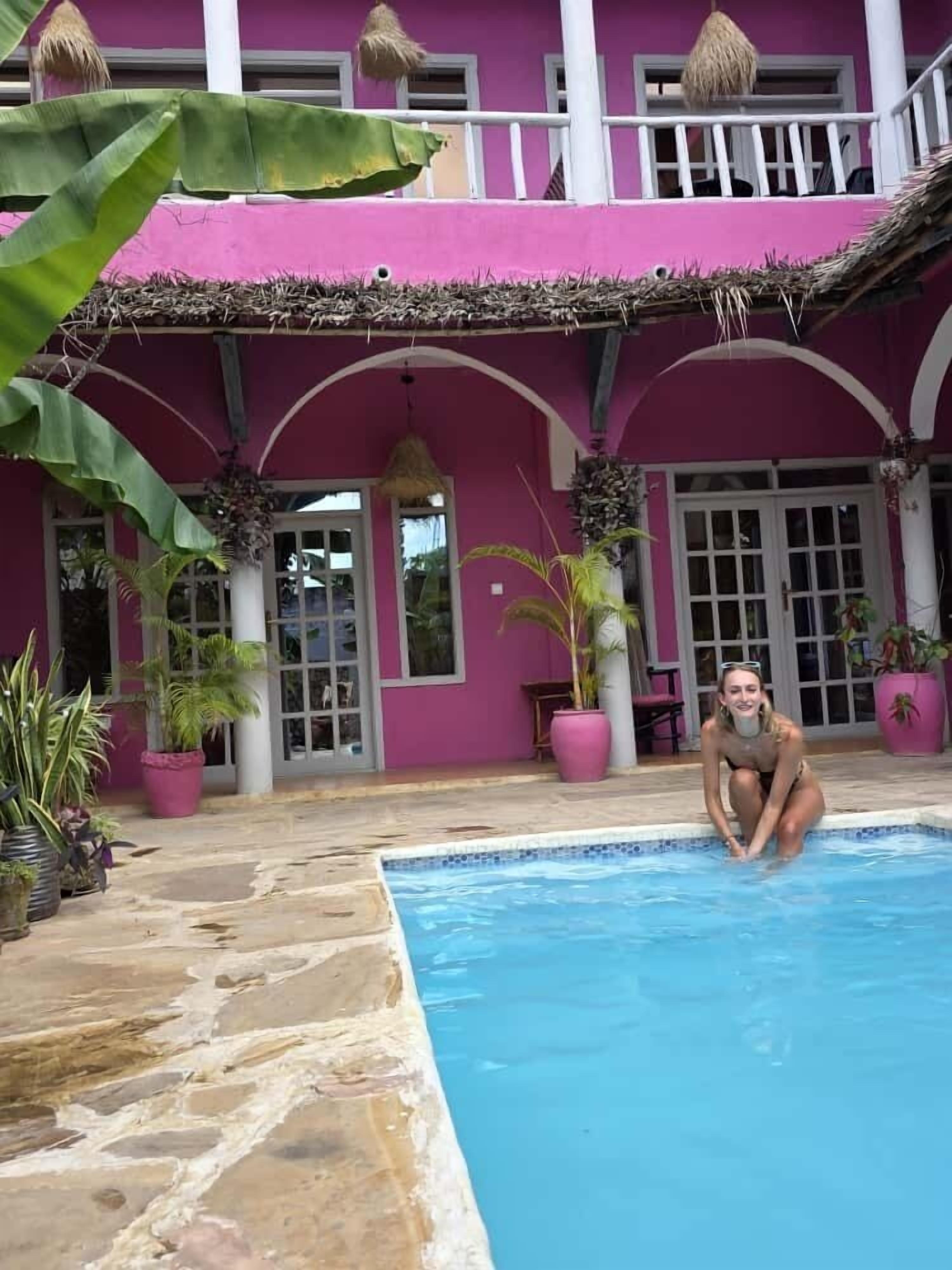 A swimming pool area with a pink building in the background, stone flooring, and potted plants.