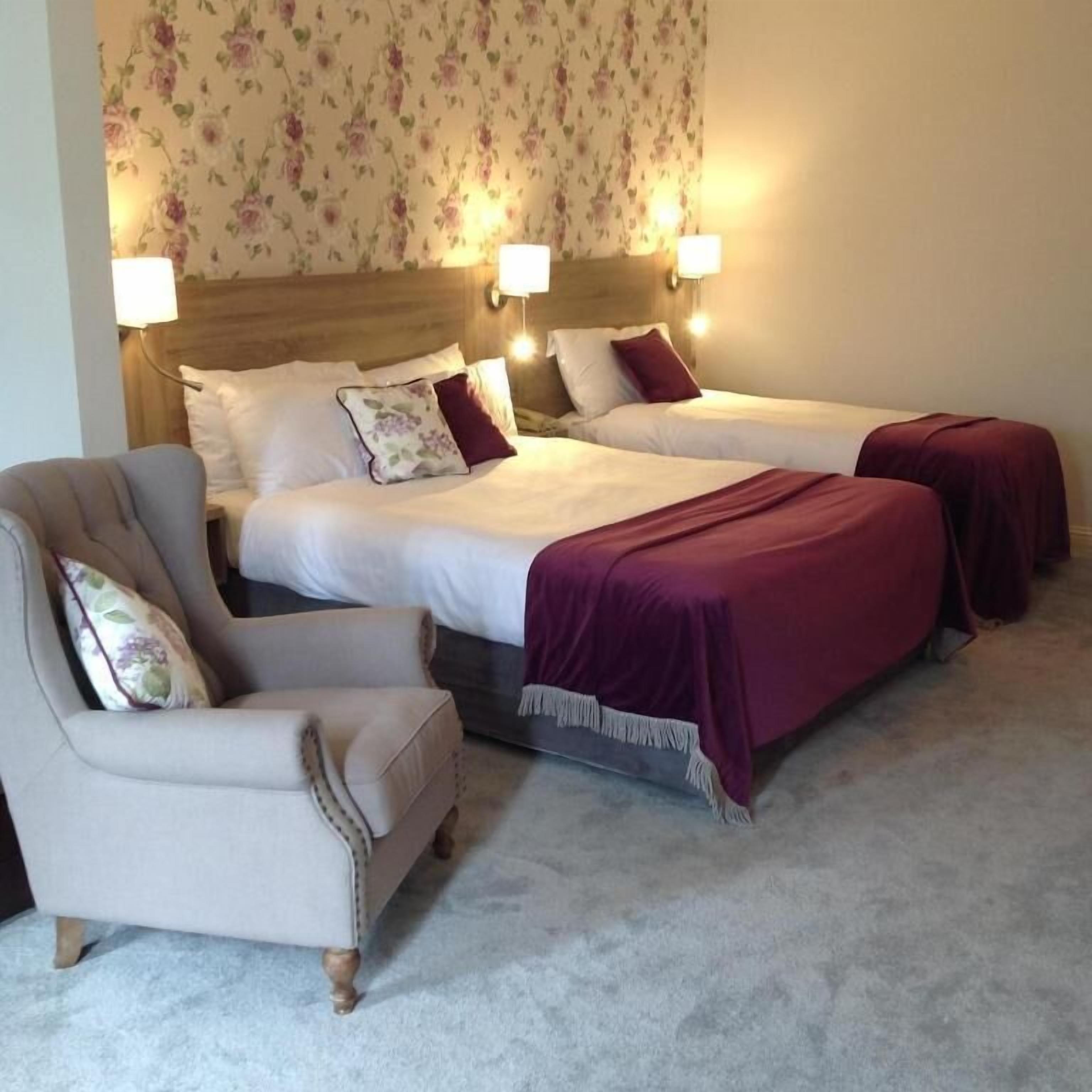 A hotel room with two beds, a chair, and floral wallpaper.
