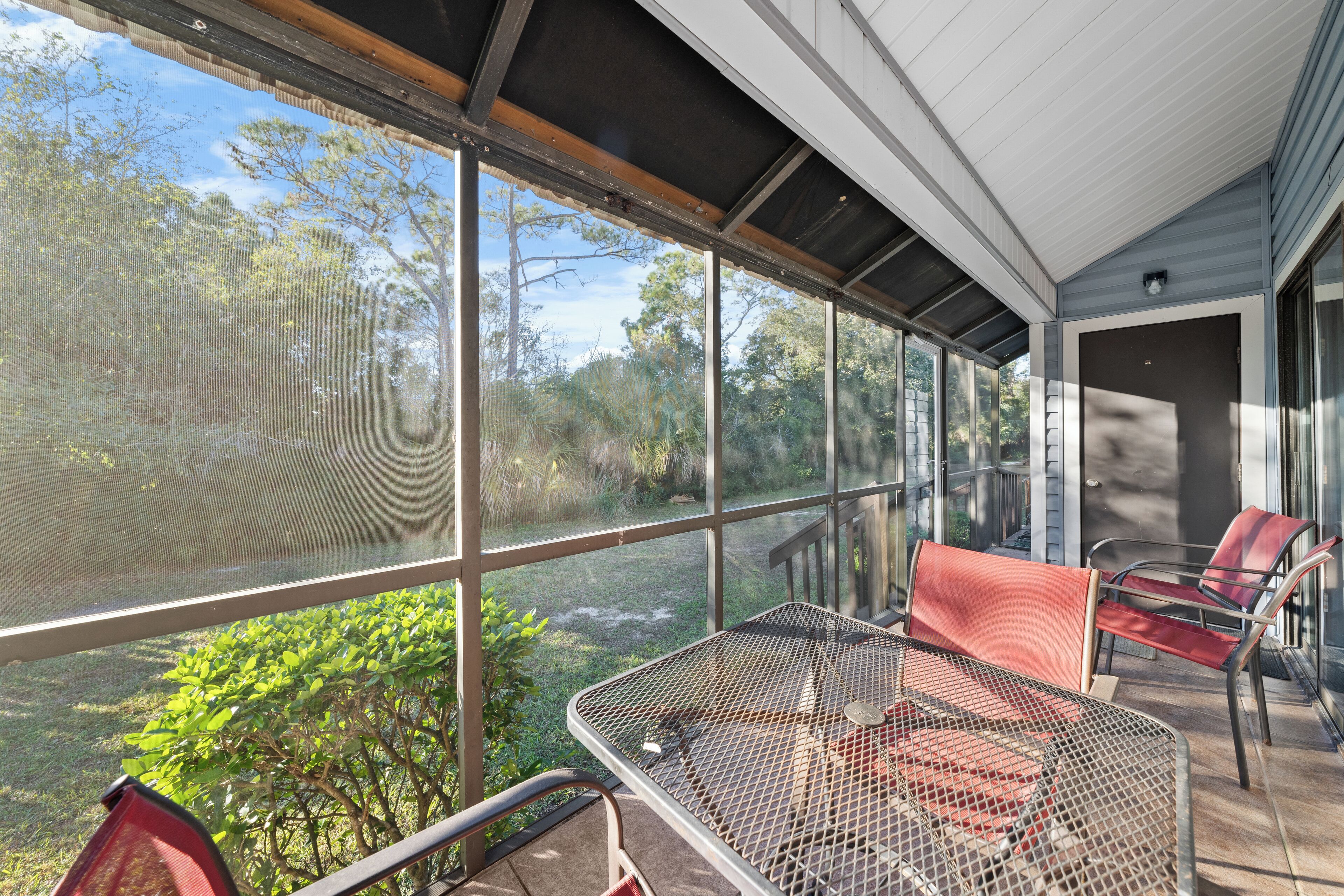 A covered outdoor area with a metal table and chairs, large windows, and a view of greenery.