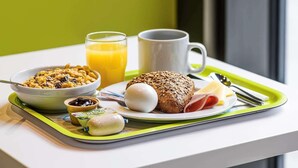 Un vassoio per la colazione con cereali, un bicchiere di succo d'arancia, una tazza, una fetta di pane, un uovo sodo, un piccolo barattolo di marmellata e una confezione di formaggio.