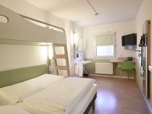Ein Hotelzimmer mit Bett, Schreibtisch mit Stuhl, Fenster, Spiegel, Fernseher und Heizkörper.
