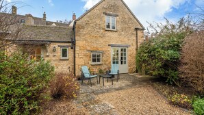 A stone house with a sloped roof, a front door, and a small garden area with two chairs and a table. - Newly Refurbished Cosy Cottage in the Heart of Chipping Campden (Chipping Campden)