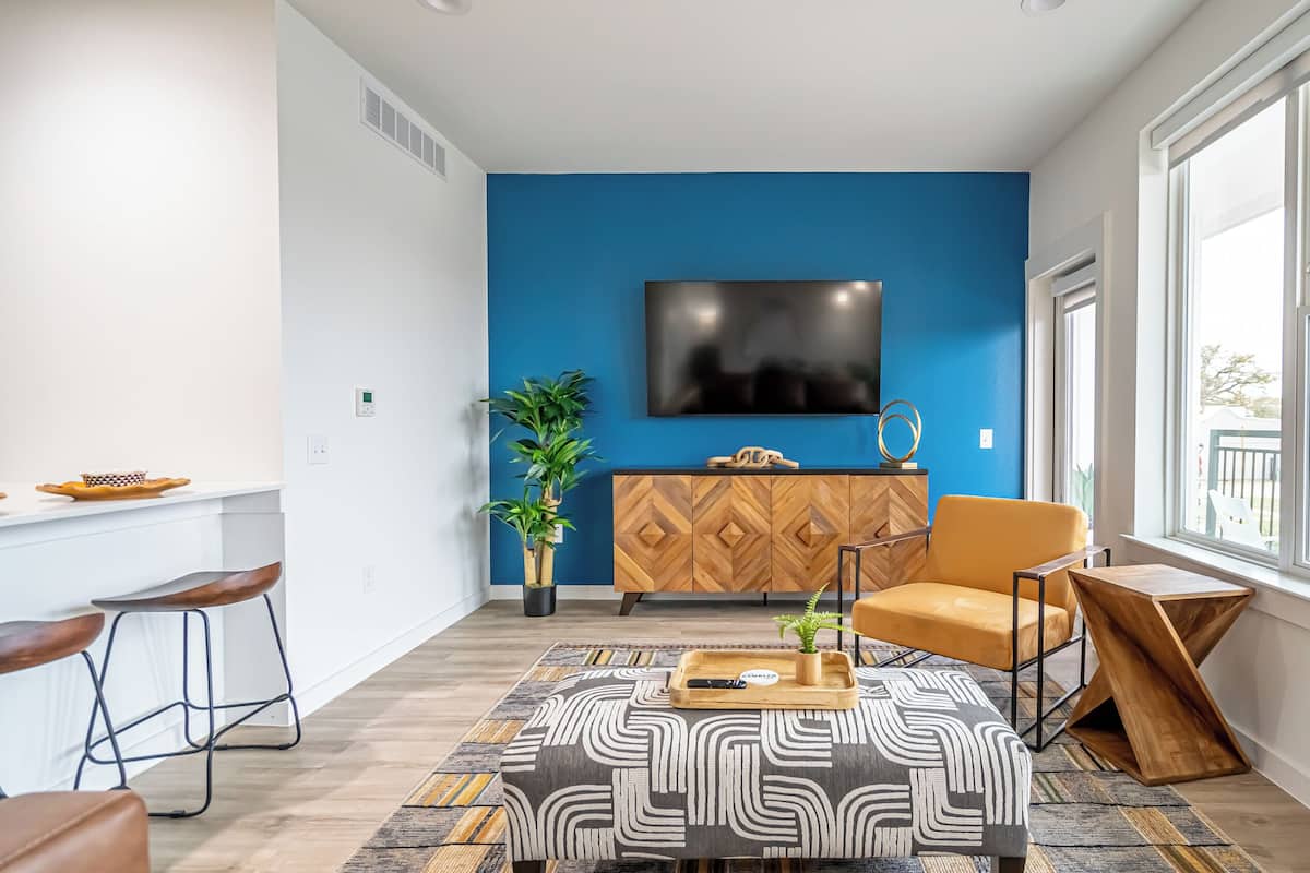 Uma sala de estar moderna com parede azul, uma televisão de tela plana, um armário de madeira e uma grande janela com vista para o exterior.