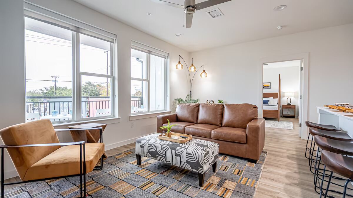 A spacious living room with a brown leather sofa, a patterned ottoman, and a modern chair. There is a large window providing natural light and a view of the outdoors.