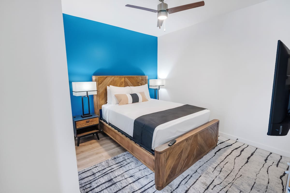 Um quarto moderno com cama de madeira, duas luminárias de apoio, um ventilador de teto e um tapete estampado.