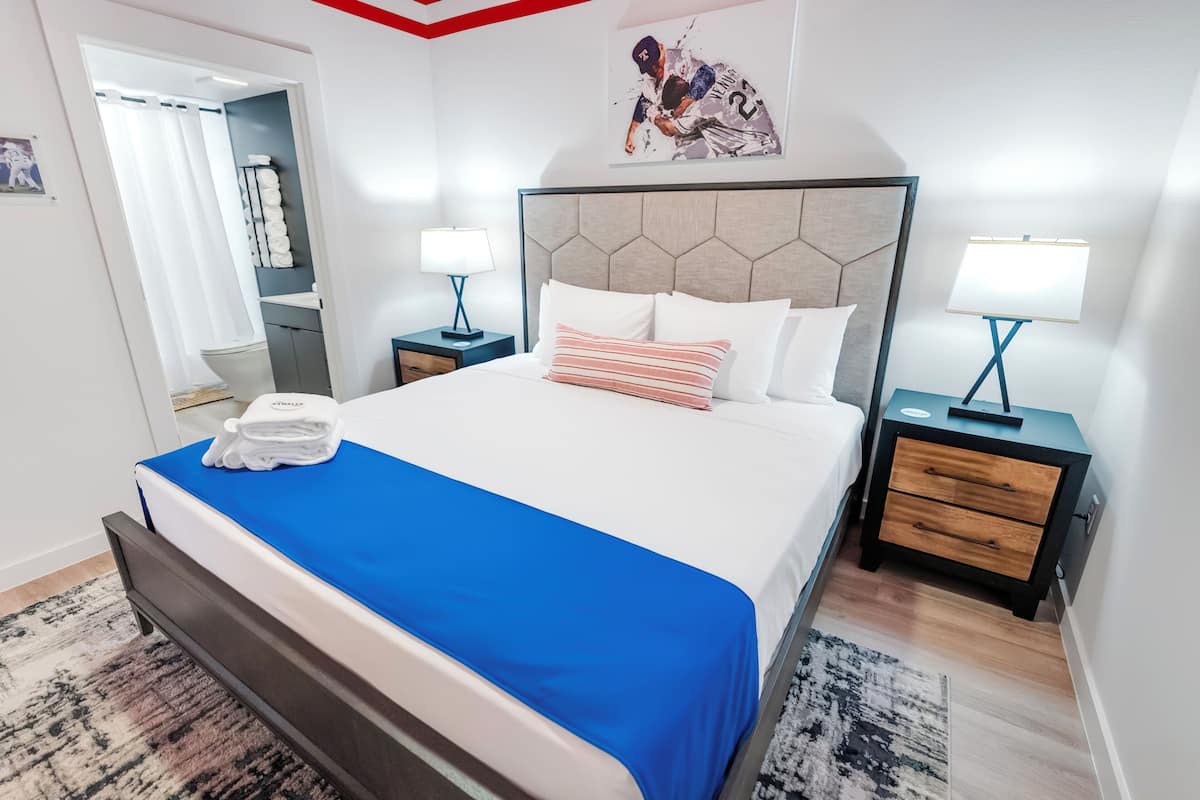 Cama bem arrumada com colcha azul, almofadas brancas e uma almofada listrada. Há duas mesas de cabeceira com abajures, um criado-mudo de madeira e um espelho na parede.