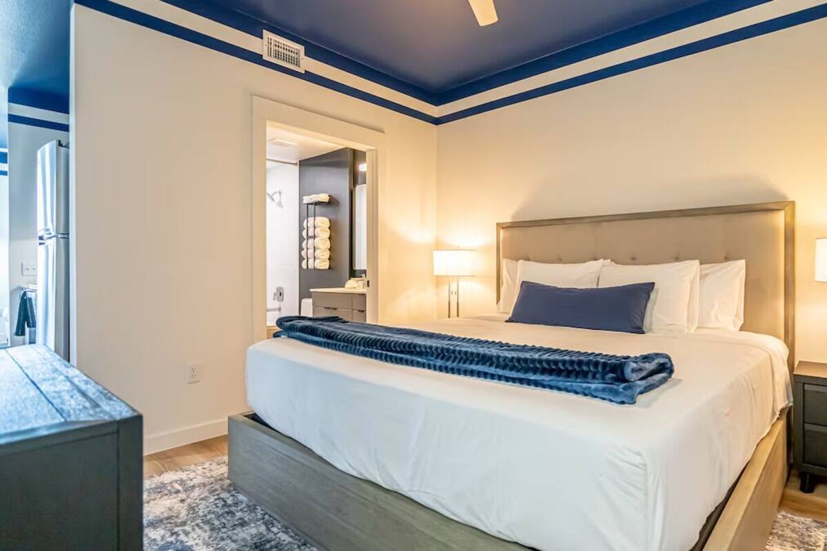 Quarto moderno com cama grande, luminárias de cabeceira e porta do banheiro visível.