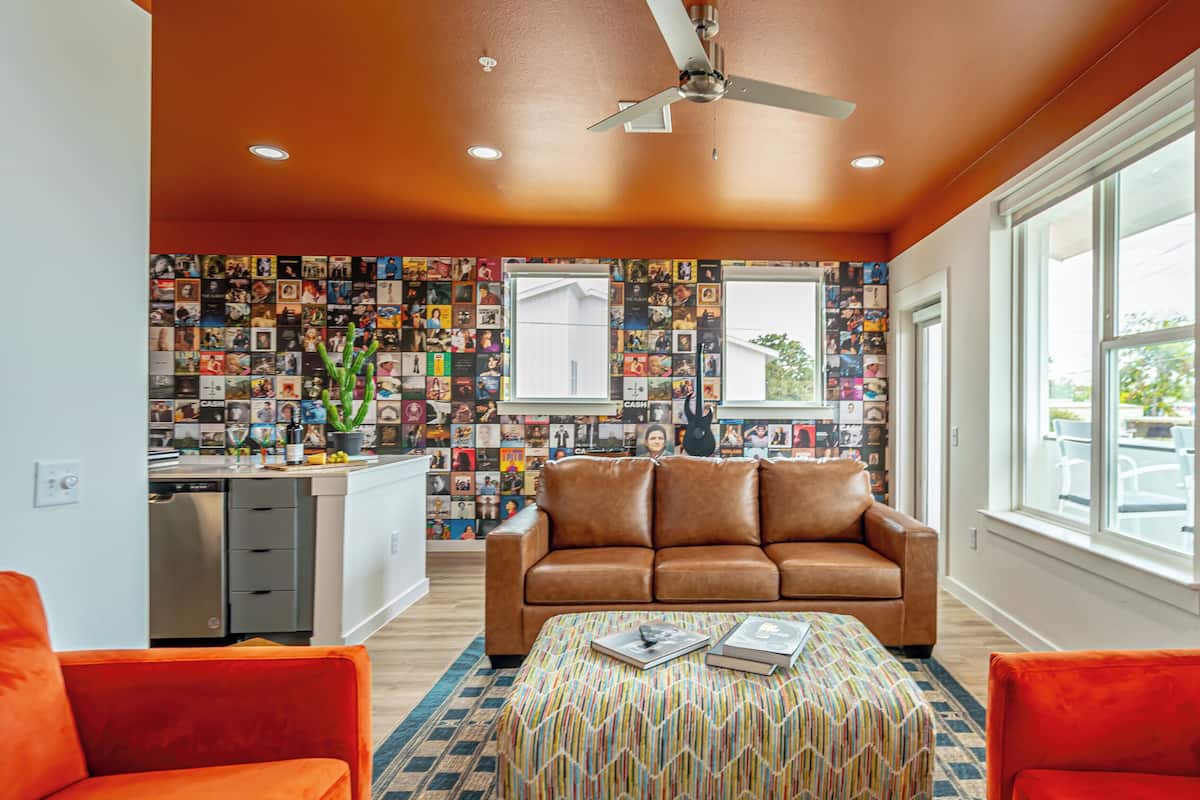 Uma sala de estar com um sofá de couro marrom, um banquinho estampado e poltronas vermelhas. É visível uma ventoinha no teto, e a parede está decorada com diversas fotografias.
